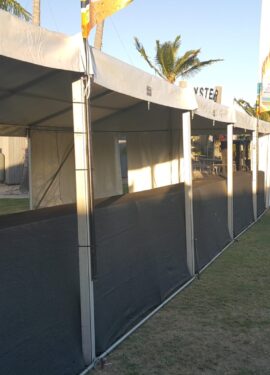Exterior side view of a long white event marquee with black mesh temporary walls on a grassy field with palm trees in the background.