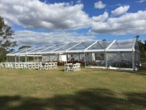 A large, open-sided marquee tent with rows of white chairs set up on a dry grassy area under a clear blue sky.