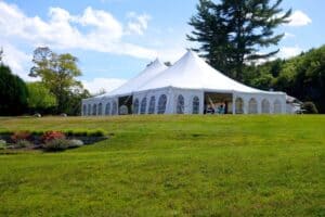 Large white twin-peak event tent set up on a manicured green lawn surrounded by trees under a sunny sky.