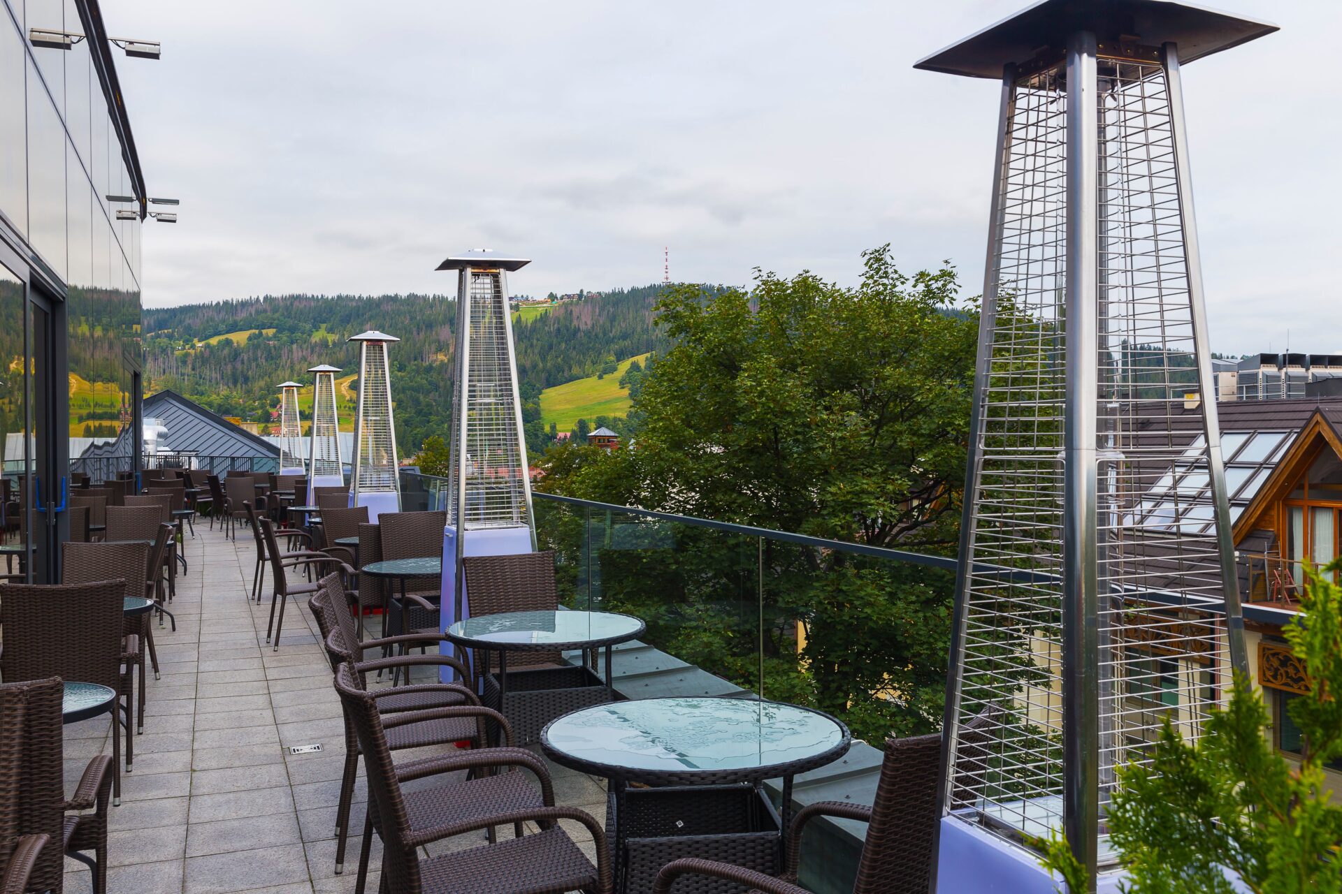 Outdoor patio seating area of a restaurant or hotel with rattan chairs, glass tables, and multiple tall pyramid gas heaters, overlooking green hills.