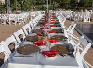 Outdoor dining table set up for an event with white chairs, gold charger plates, and red napkins.