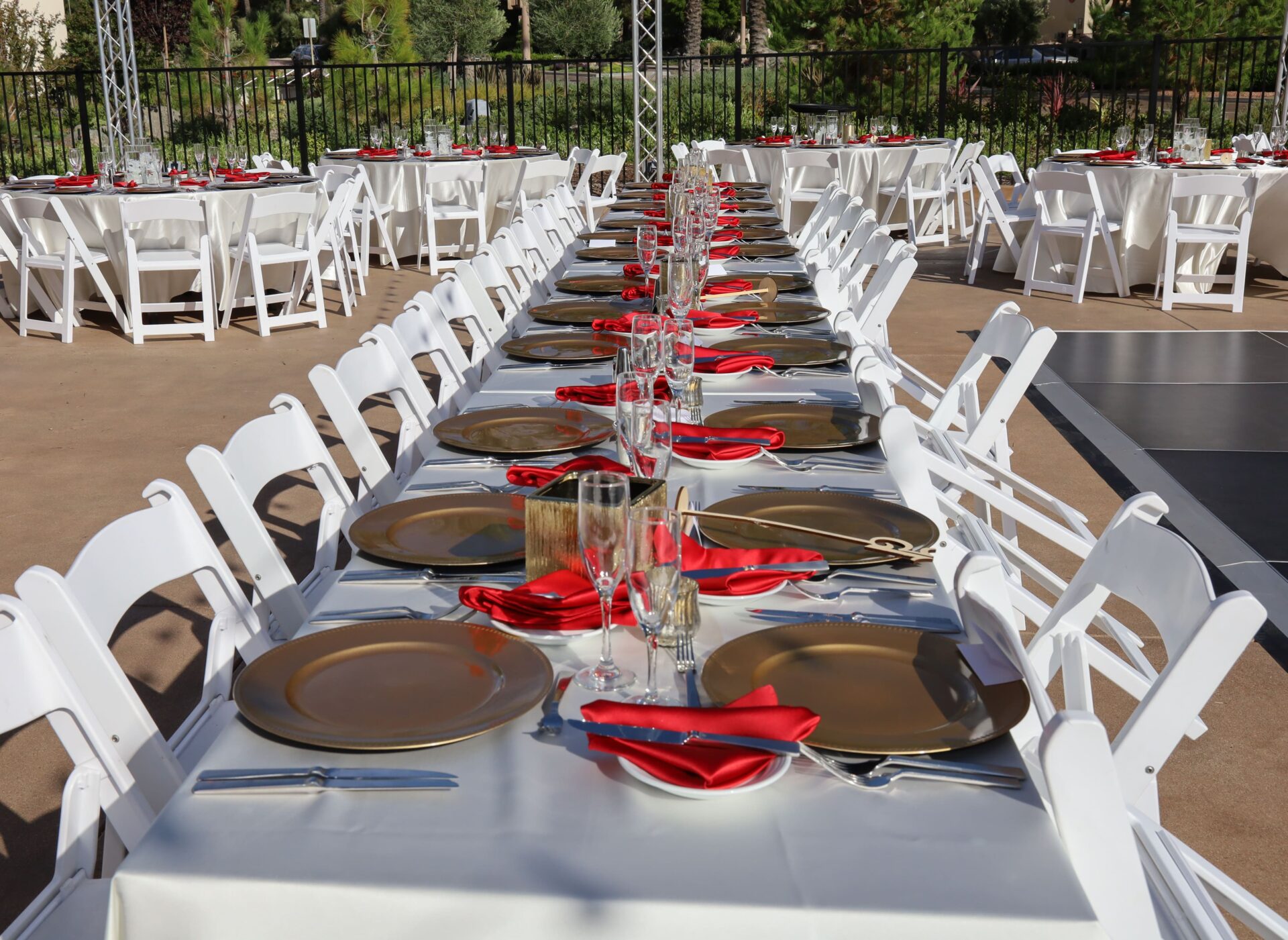 Outdoor dining table set up for an event with white chairs, gold charger plates, and red napkins.