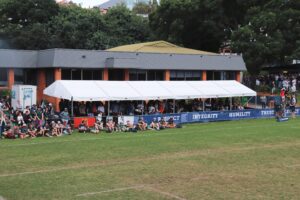 A white tent shelters spectators sitting on the grass at an outdoor event.