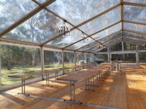 Interior view of a clear span marquee tent with long wooden tables and black chandeliers hanging from the transparent ceiling.