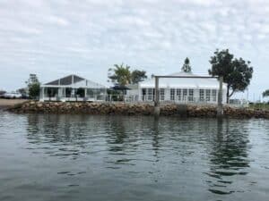 A white marquee tent and a white building set up on a rocky shore next to the water.