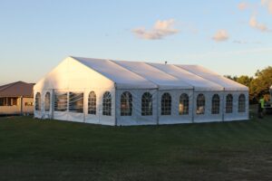 A large white marquee tent with arched windows set up on a grassy area under a partly cloudy sky.