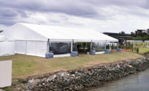 An aluminum frame marquee tent set up outdoors on a grassy area near water.