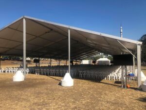 A large, open-sided marquee tent with rows of white chairs set up on a dry grassy area under a clear blue sky.