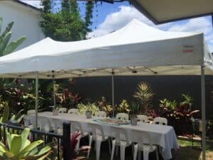 A white pop-up canopy tent covering a long table with white chairs in a backyard setting.