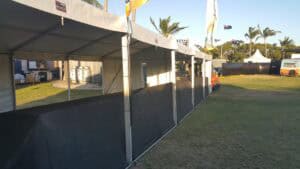 Exterior side view of a long white event marquee with black mesh temporary walls on a grassy field with palm trees in the background.