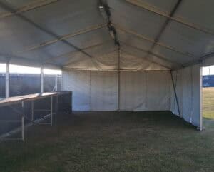 Interior view of a large temporary event marquee or tent with a metal frame, fabric walls, and a dirt floor with patches of grass.